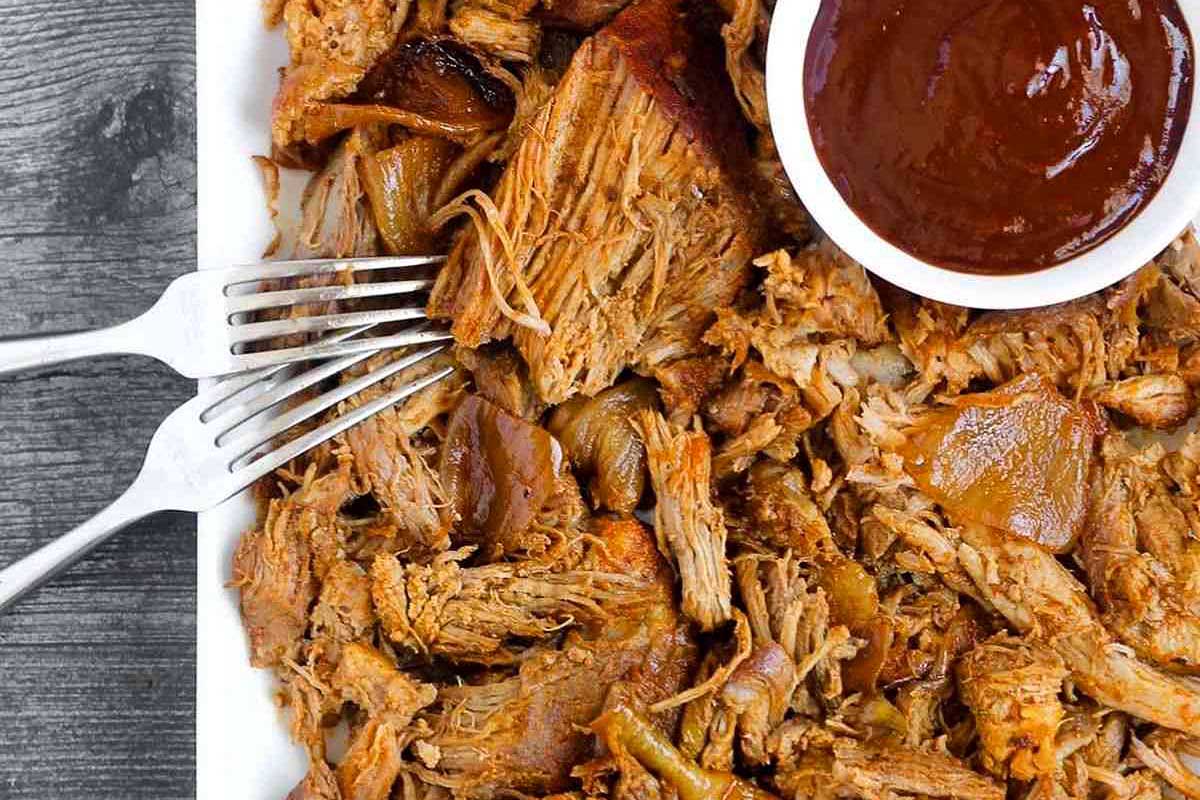 A plate of slow-cooked pulled pork with caramelized onions and a side of barbecue sauce, served with two forks.
