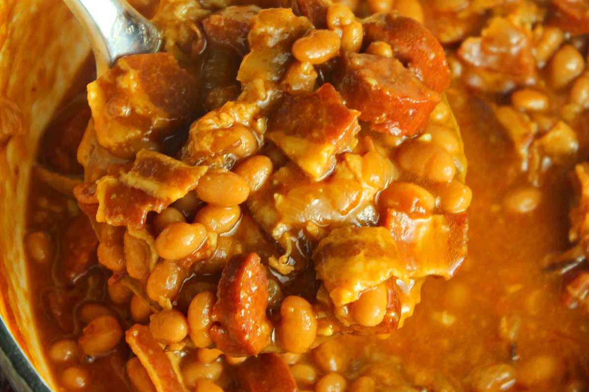 A close-up of a spoon scooping baked beans and chunks of pork from a smoker recipe, focusing on the texture and saucy consistency.