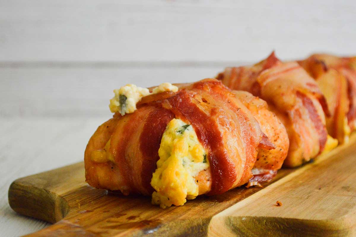 A smoker recipes-inspired bacon-wrapped stuffed chicken breast on a wooden cutting board, with melted cheese oozing out, set against a white wooden background.