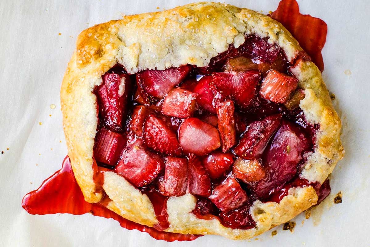 Freshly baked strawberry rhubarb galette from our top rhubarb recipes, on a white surface, with juices spilling from the crust.