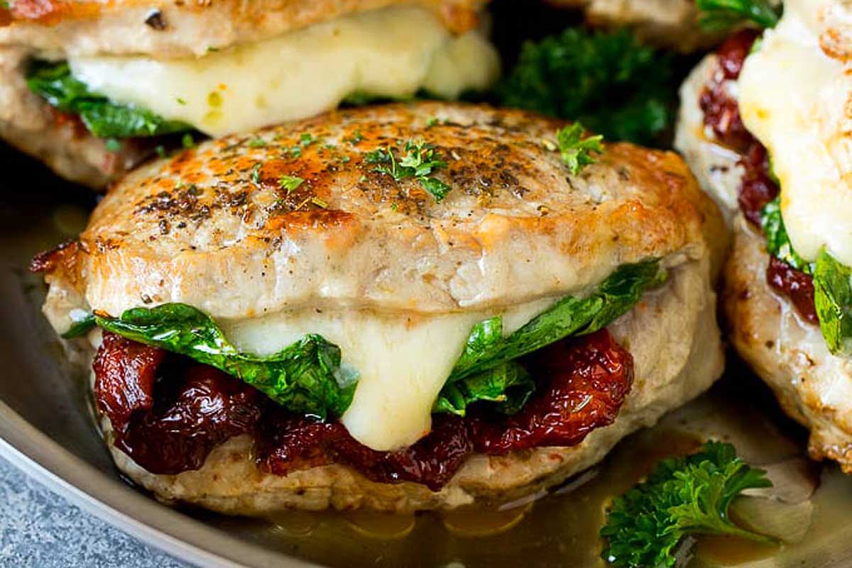 A close-up of a pork chop sandwich with melted cheese, sundried tomatoes, and spinach on a herb-crusted bun, displayed on a plate.