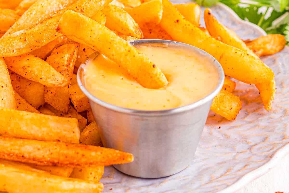 A plate of seasoned french fries with a small metal cup of Taco Bell copycat dipping sauce, garnished with parsley, on a white surface.