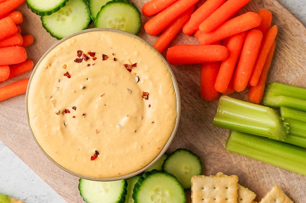 A bowl of creamy Velveeta cheese dip surrounded by sliced cucumbers, carrots, celery, and crackers on a wooden cutting board.