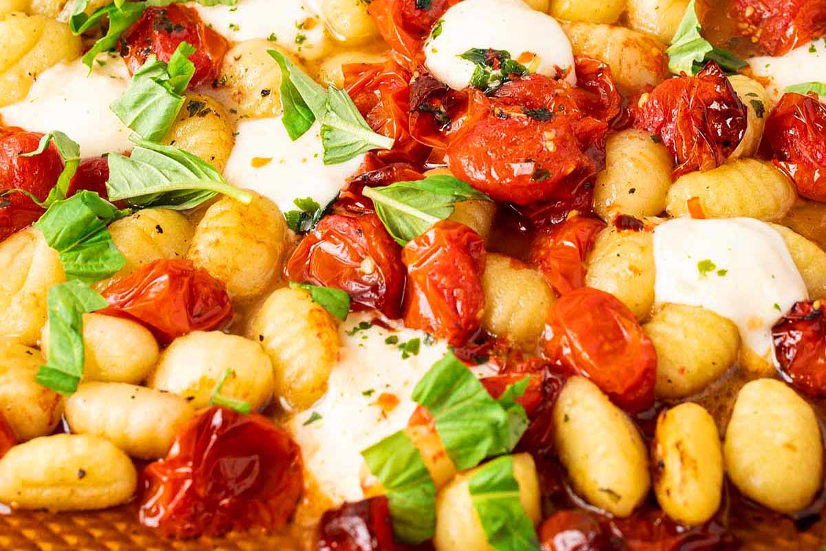 Close-up image of a sheet pan recipe featuring cooked gnocchi with roasted cherry tomatoes, fresh basil, and melted cheese.