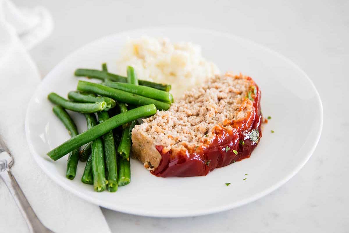 A plate of meatloaf with ketchup glaze from our ketchup recipes, served with green beans and mashed potatoes on a white background.