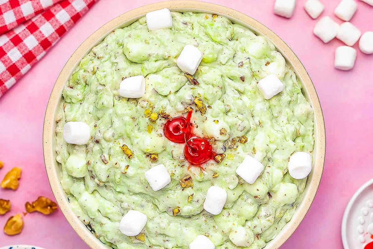 A bowl of green pistachio pudding mix ambrosia salad garnished with marshmallows and a cherry on top.