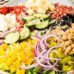 A colorful chopped Italian salad platter featuring diced yellow bell peppers, red onion slices, chickpeas, black olives, cherry tomatoes, cucumber slices, fresh mozzarella, and sliced meat next to wooden salad tongs.