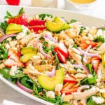 A vibrant Strawberry Fields salad, featuring spinach, sliced strawberries, avocado, red onions, grilled chicken, feta cheese, and almonds. Served in an oval white dish with a side of tangy dressing and chopped nuts for added crunch.