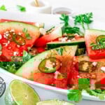 A platter of spicy watermelon jalapeño salad garnished with herbs and feta cheese. A bowl of crumbled feta, lime halves, and fresh herbs surround the platter.
