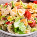 A bowl of BLT pasta salad with farfalle pasta, chopped lettuce, tomatoes, cheddar cheese cubes, and crispy bits of bacon, all lightly dressed with a creamy dressing.