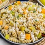 A bowl of dill pickle pasta salad with creamy dressing, garnished with chopped dill, and mixed with cubes of cheese and pickles on a woven placemat.