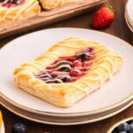 A rectangular cream cheese berry Danish with a fruit filling and icing drizzle sits on a white plate. Surrounding it are more pastries, whole strawberries, blueberries, and sliced oranges on a wooden table.