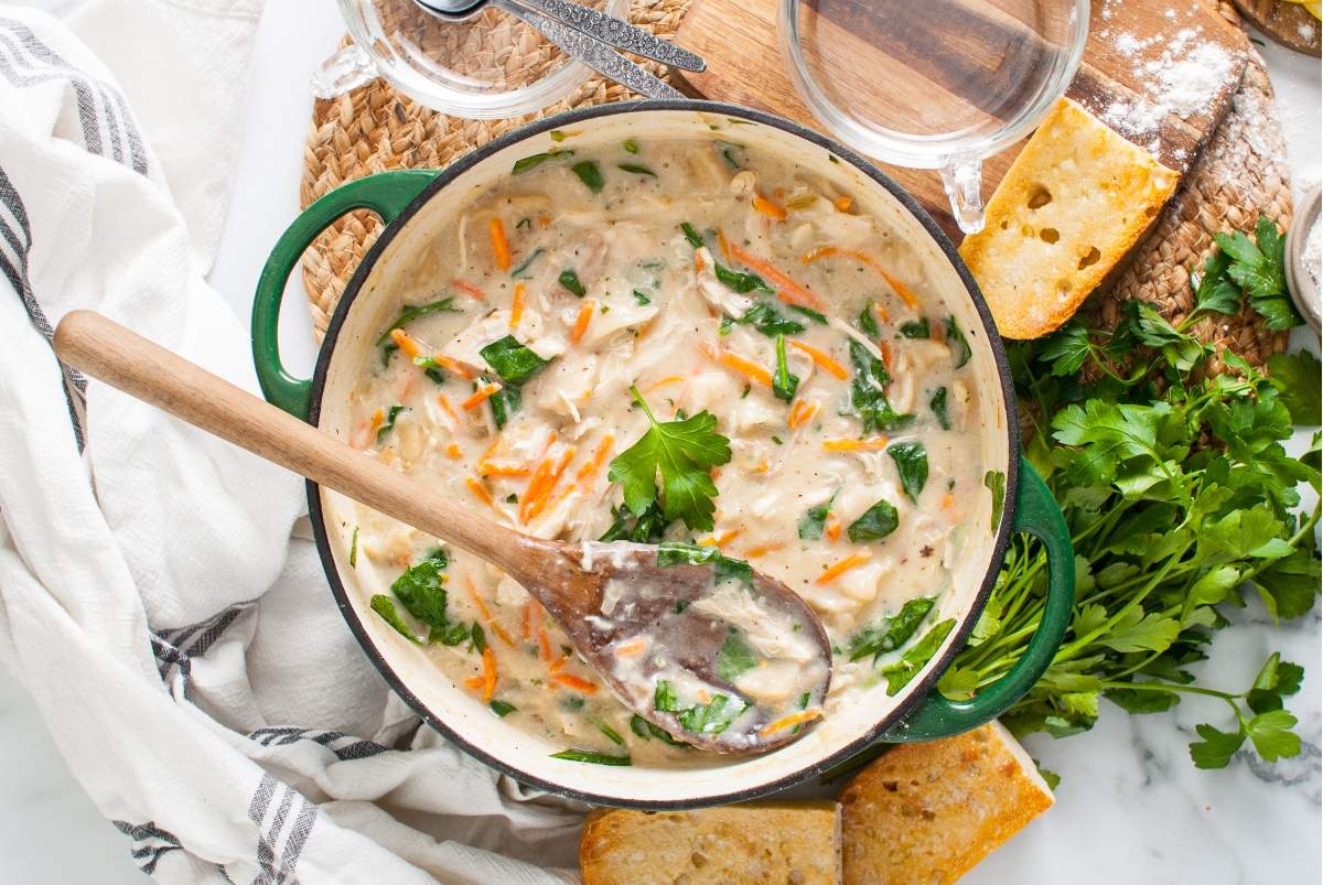 A creamy chicken, spinach, and carrot soup in a green pot, garnished with parsley. A wooden spoon rests inside the pot. Surrounding the pot are pieces of toasted bread, fresh parsley, a cutting board, and a white cloth.