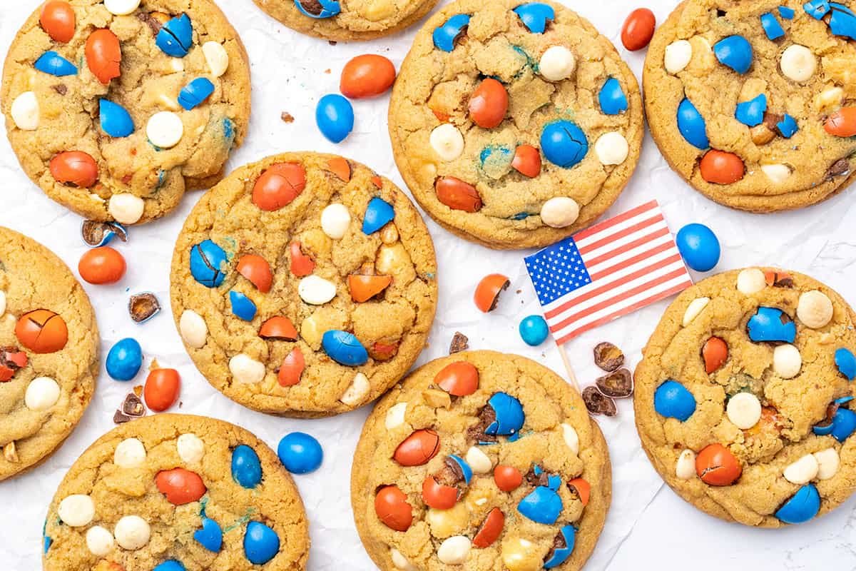 A batch of patriotic desserts featuring cookies with red, white, and blue candies is arranged on a white surface alongside a small American flag.