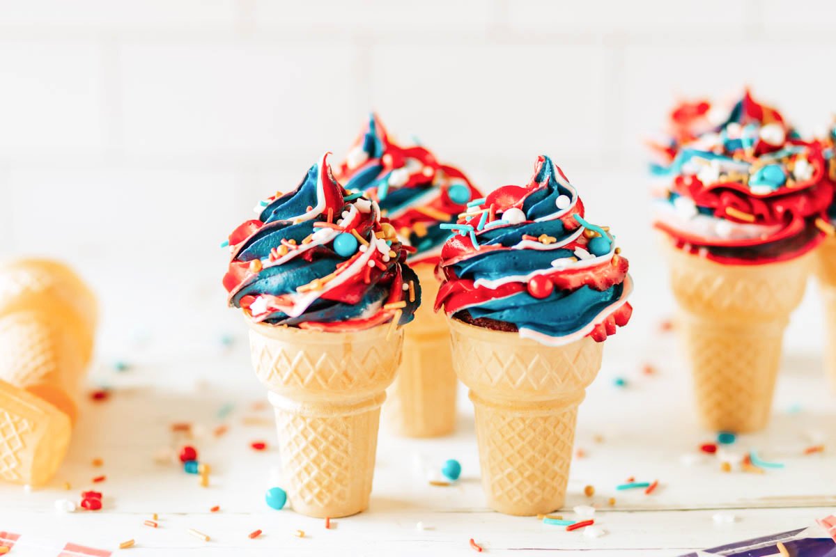 Patriotic desserts featuring ice cream cones with colorful blue, red, and white swirled frosting and sprinkles on top, placed on a white surface with scattered sprinkles.