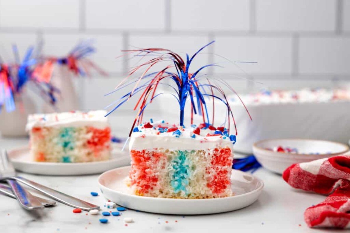 A slice of red, white, and blue cake with frosting and sprinkles, topped with metallic blue and red decorations, sits on a white plate. Other cake slices and utensils are visible in the background, showcasing an array of patriotic desserts.