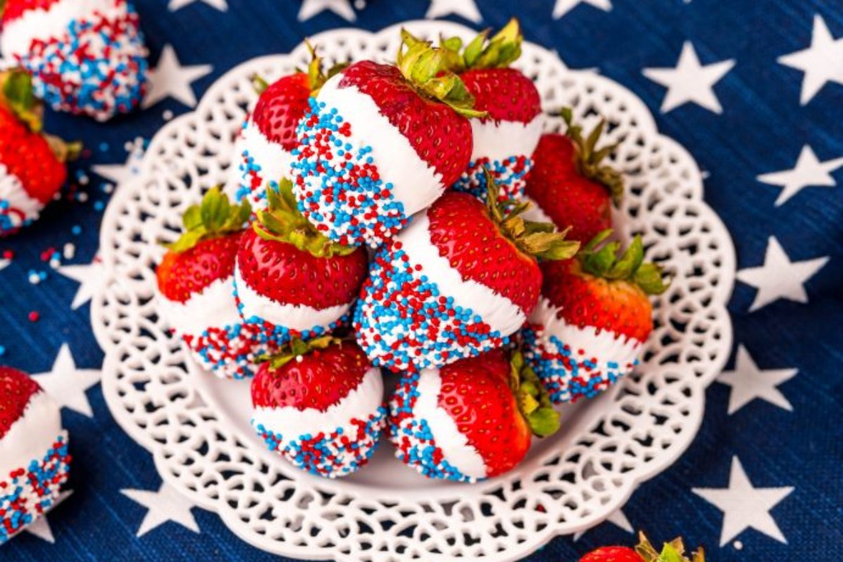 Patriotic desserts, like strawberries dipped in white chocolate and decorated with red, white, and blue sprinkles, are arranged on a white lace-patterned plate, placed on a blue cloth with white star designs.