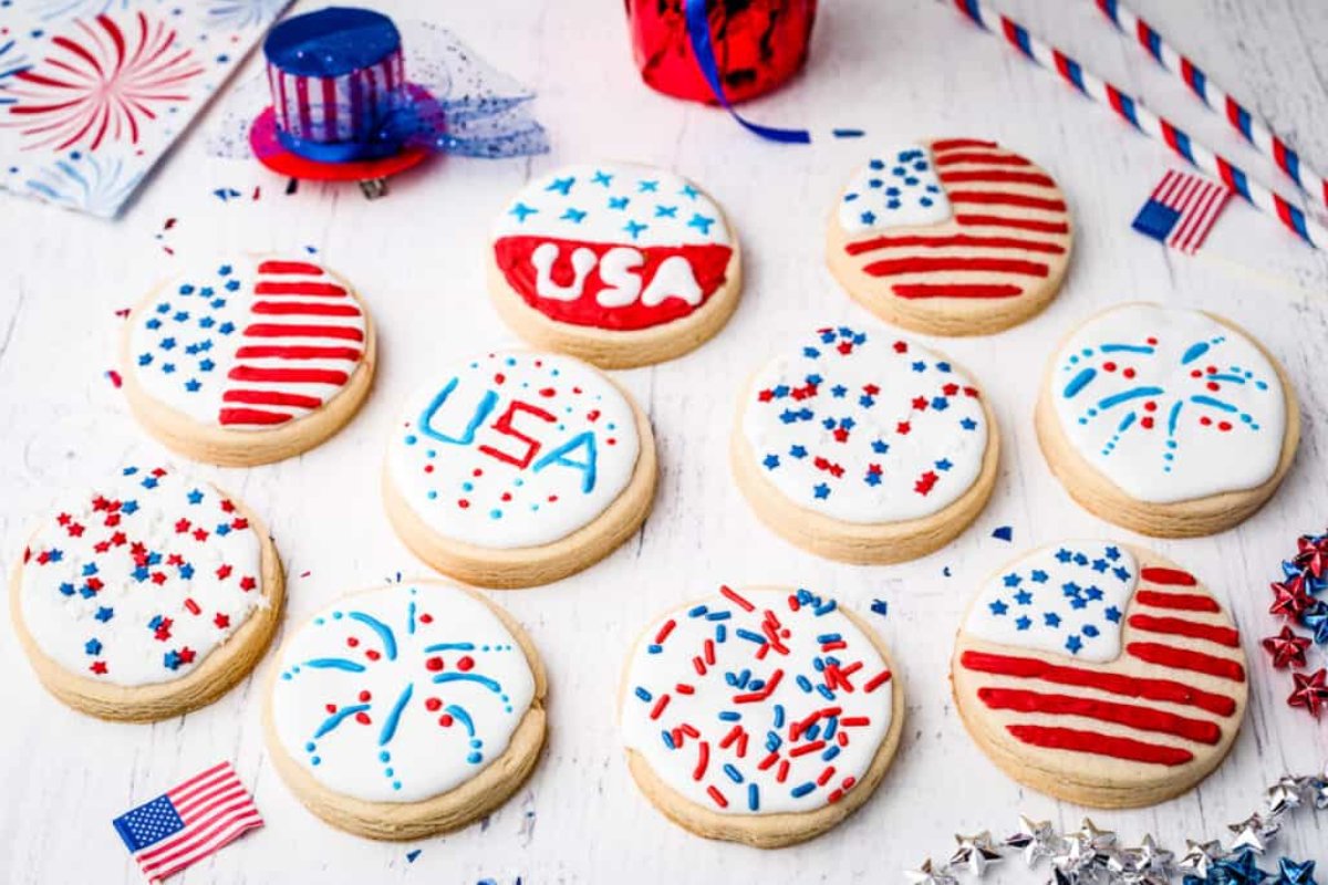 A selection of frosted cookies decorated with American flag themes, fireworks, and "USA" in red, white, and blue colors, placed on a white surface with patriotic decorations in the background. These patriotic desserts are perfect for celebrating Independence Day in style.