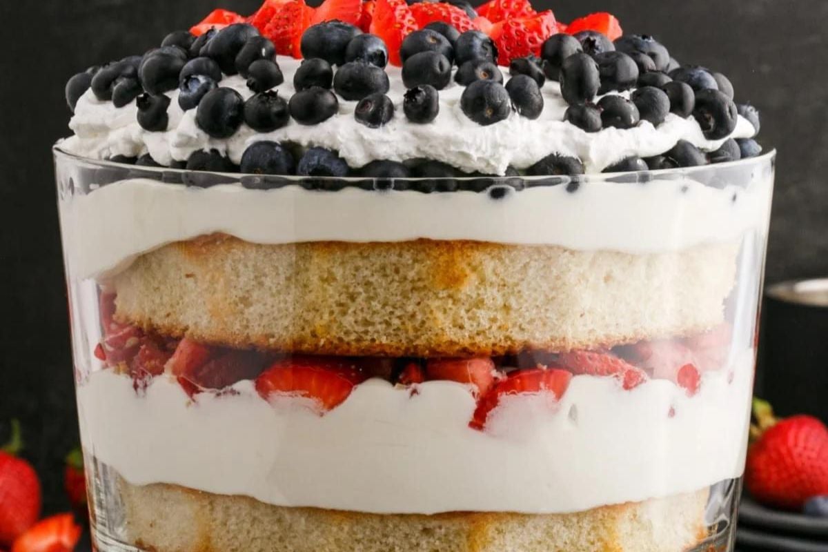 A layered dessert in a glass bowl with cool whip, blueberries, strawberries, sponge cake, and cream filling.