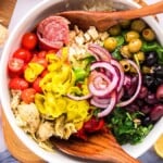 A colorful antipasto salad with assorted vegetables, meats, and olives, served in a white bowl with wooden utensils.