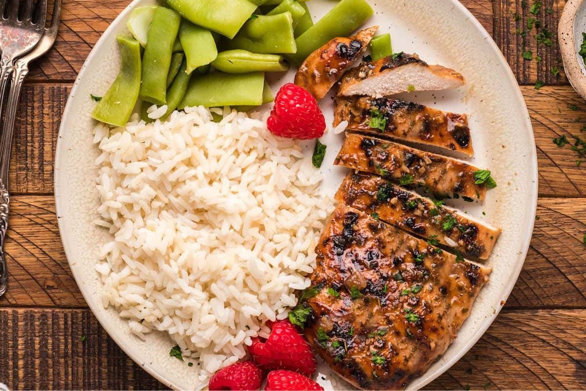 A delicious plate featuring one of our favorite chicken recipes: grilled chicken served with white rice, green beans, and raspberries on a wooden table.