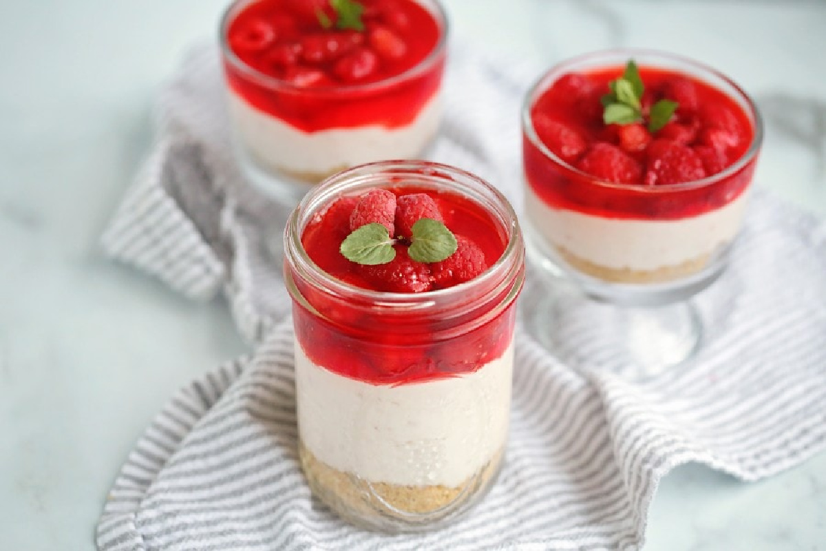 Three jars of creamy layered dessert with a biscuit base, topped with red jelly and fresh raspberries, garnished with mint leaves on a striped cloth.