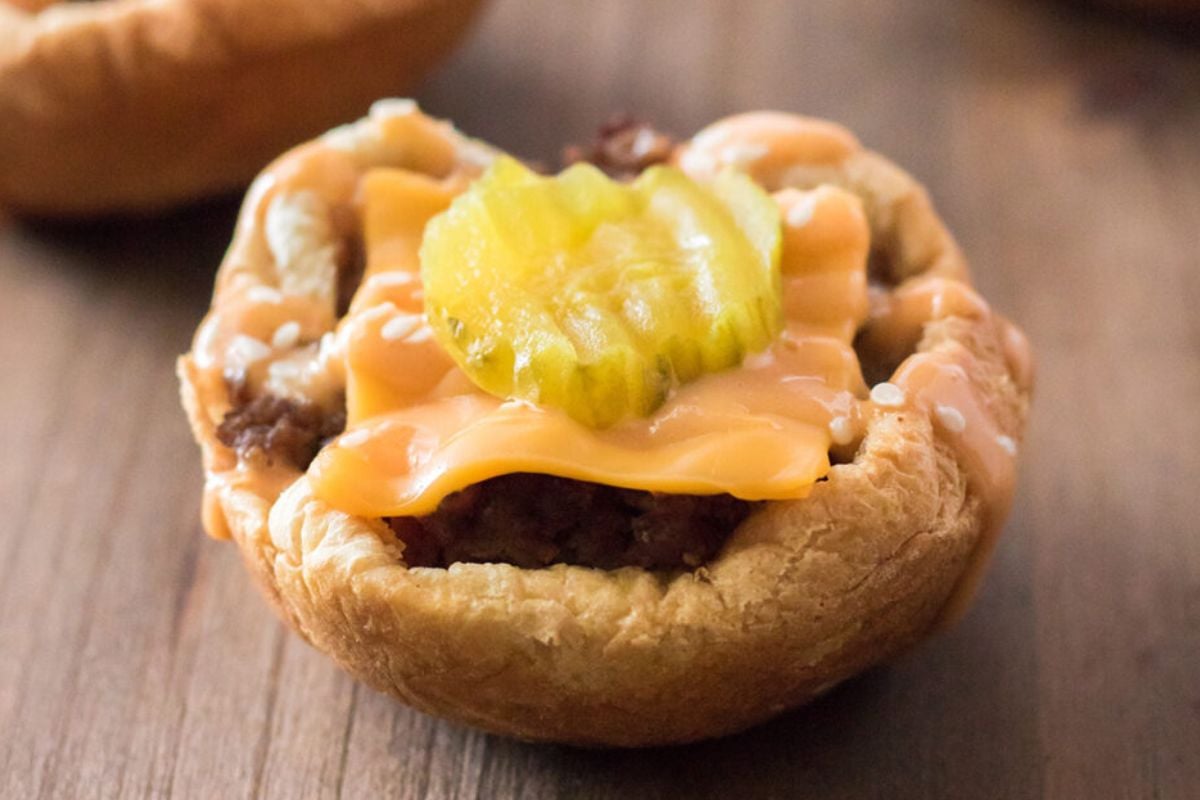 A small bread cup filled with ground meat, topped with melted cheese, and a pickle slice, placed on a wooden surface—perfect for those seeking Big Mac-inspired recipes.