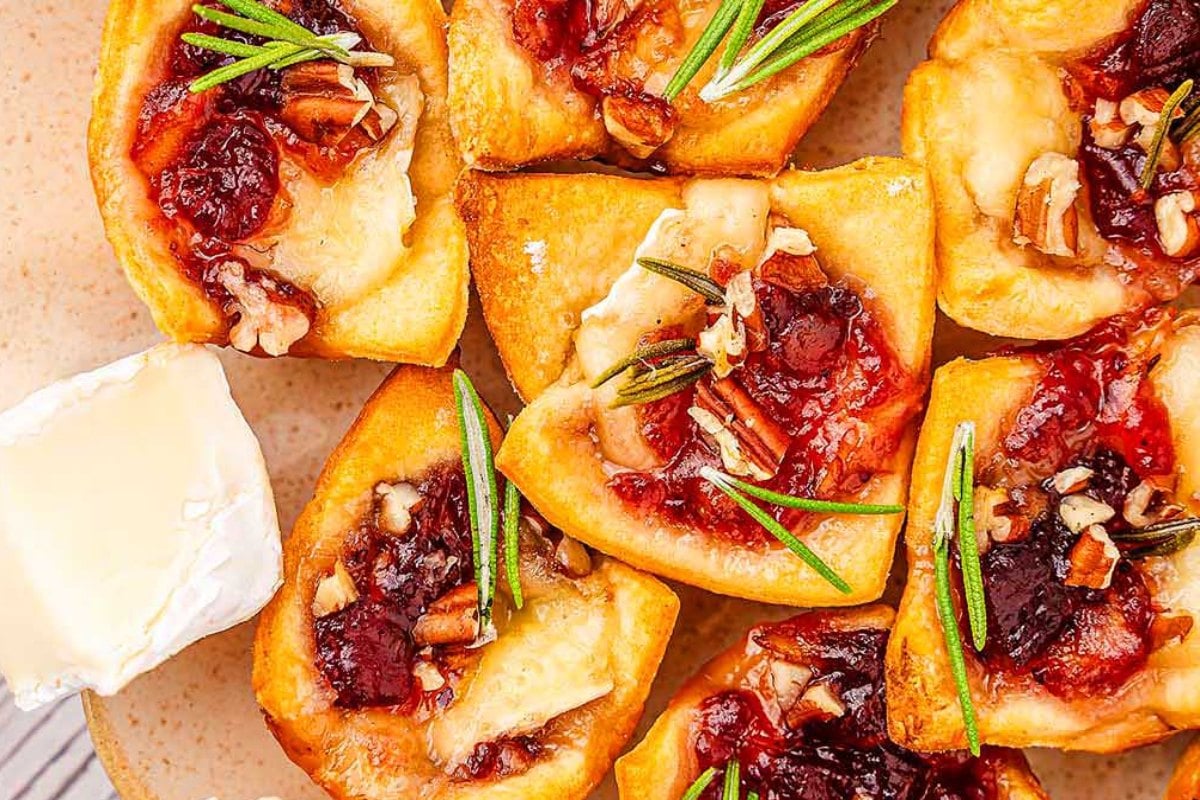 Golden pastry bites made from crescent roll recipes, topped with brie, cranberry sauce, pecans, and rosemary, arranged on a plate.