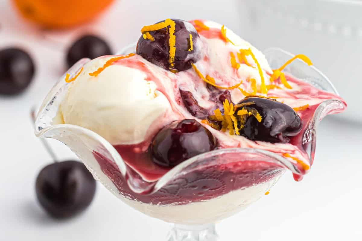 A glass bowl holds vanilla ice cream topped with dark cherries and drizzled with cherry sauce, garnished with orange zest. Cherries are scattered in the background, making it a delightful addition to any cherry recipes collection.