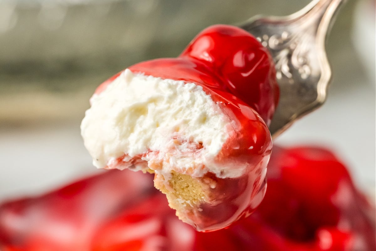 A close-up of a fork holding a bite of a dessert with a layer of whipped cream and a bright red topping, possibly strawberry or cherry, over a base that appears to be graham cracker crust.