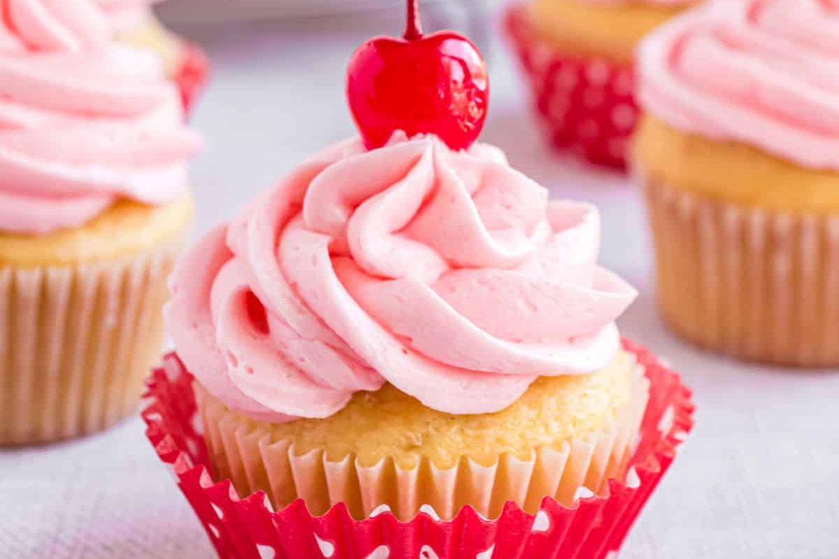 A vanilla cupcake with pink frosting topped with a cherry, placed in a red polka-dotted cupcake liner, showcasing one of the delightful treats from our favorite cherry recipes. Other similarly decorated cupcakes are visible in the background.