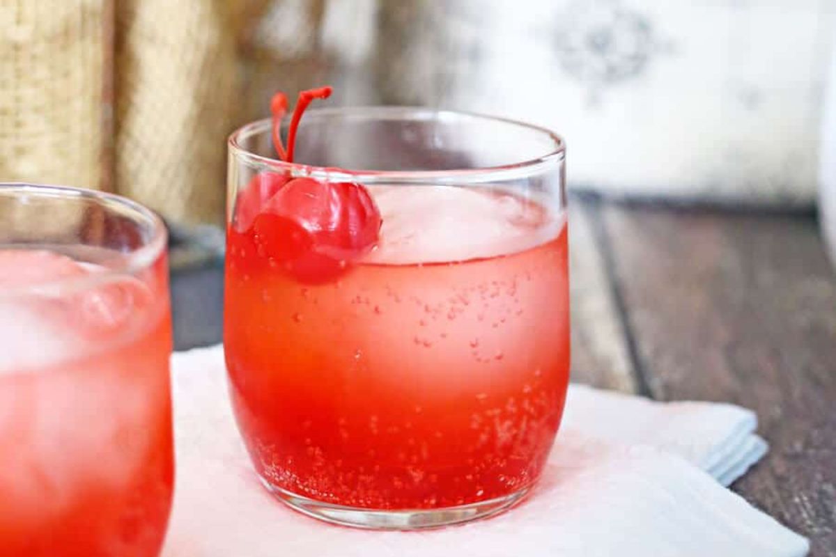 A glass of red beverage with ice and maraschino cherries on top, placed on a white napkin on a wooden table, perfect for showcasing in cherry recipes.