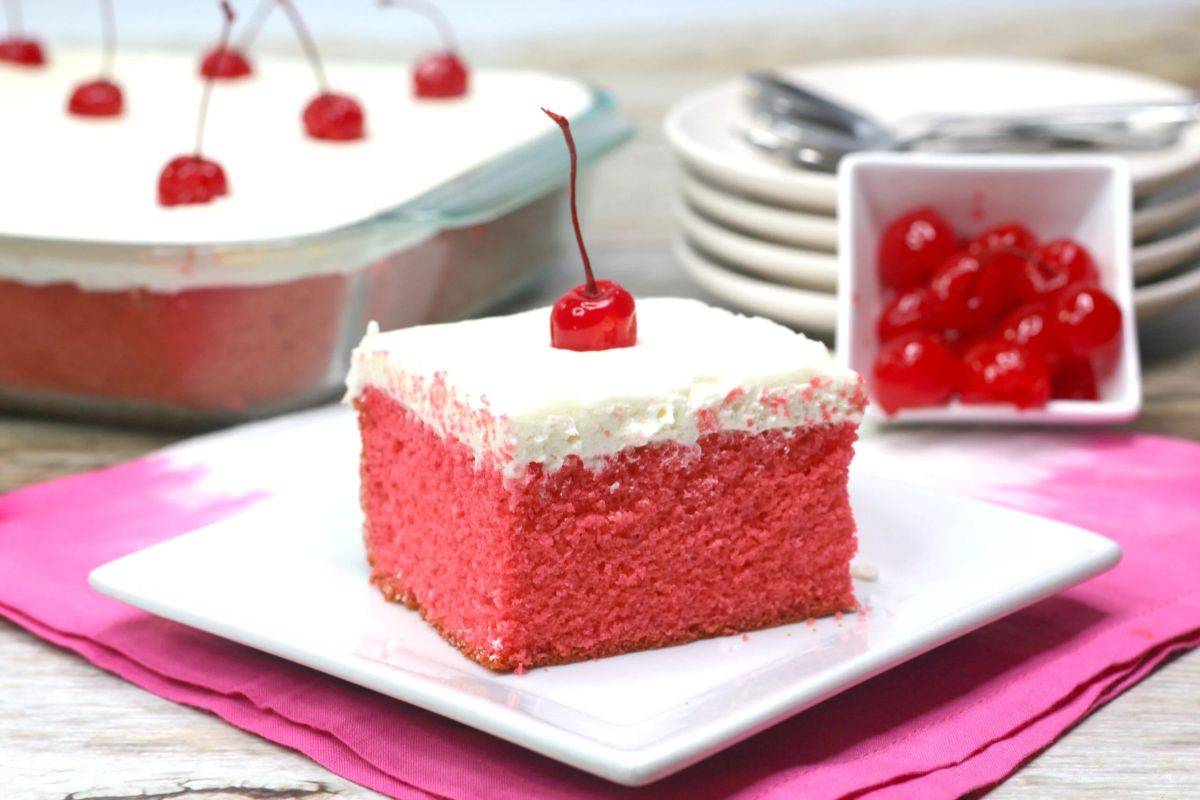 A slice of pink cake with white frosting topped with a cherry, on a white plate. A larger portion of the cake and a bowl of cherries are in the background, perfect inspirations for cherry recipes. Plates and forks are stacked beside them.
