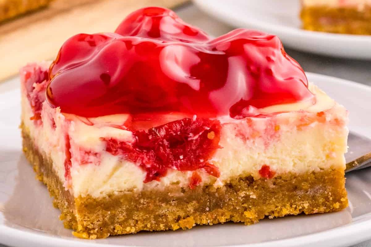A slice of cheesecake bar topped with a glossy cherry compote, featuring a graham cracker crust and served on a white plate—perfect for those who love cherry recipes.