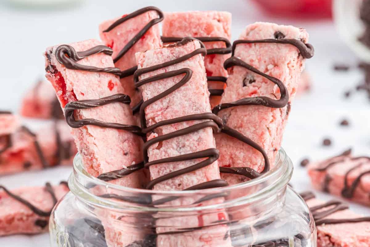 A jar filled with rectangular pink cookies drizzled with chocolate, inspired by delightful cherry recipes, with more cookies scattered around.