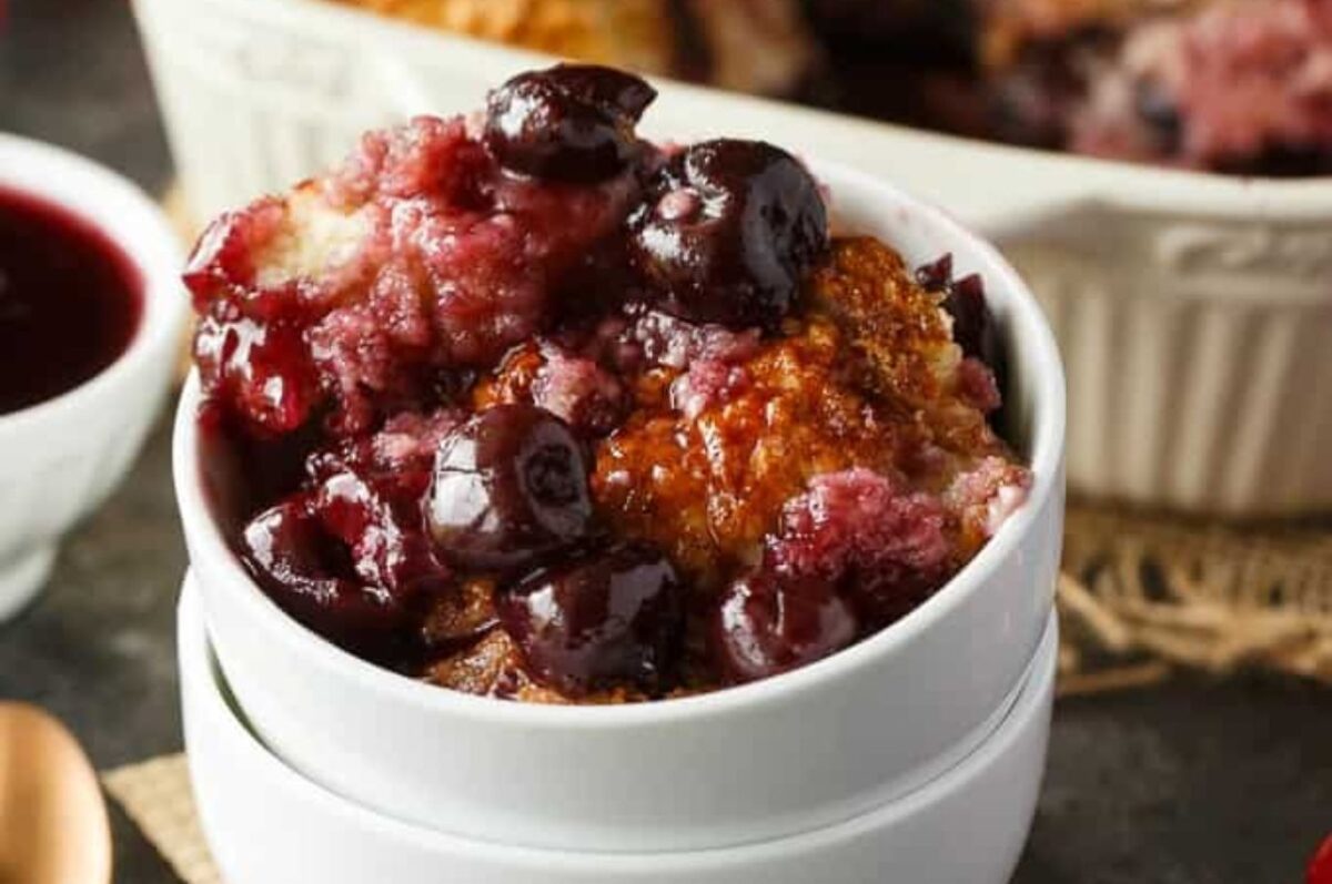 A close-up of a bowl filled with cherry cobbler, featuring baked golden-brown dough and juicy dark cherries. In the background is a smaller bowl of cherry sauce, showcasing one of the best cherry recipes for dessert lovers.