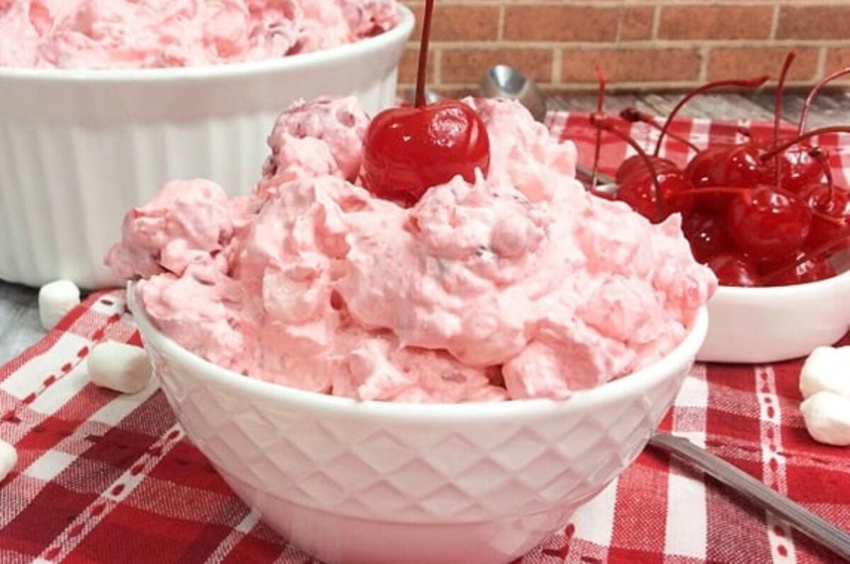A bowl of pink cherry fluff dessert topped with a cherry, placed on a red and white checkered cloth. A larger dish of the same dessert and a bunch of cherries are in the background, showcasing one of the most delightful cherry recipes you'll ever try.
