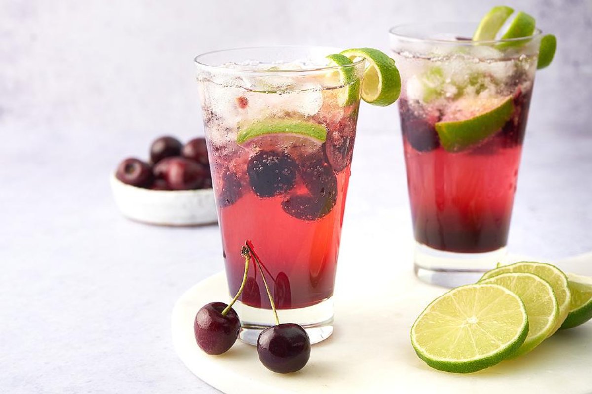 Two glasses of cherry limeade with ice, garnished with lime slices, with fresh cherries and lime slices on the side. A bowl of cherries is in the background, showcasing one of the best cherry recipes for a refreshing drink.