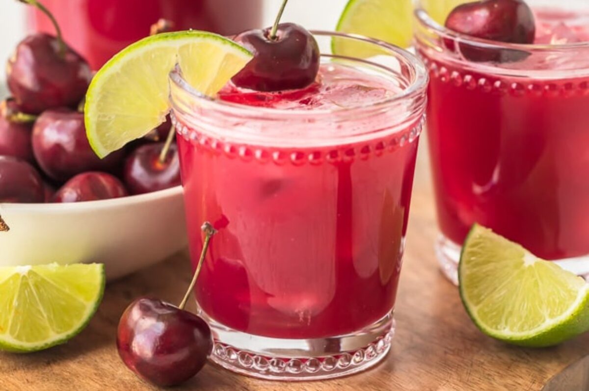 Two glasses of red cherry limeade, garnished with cherries and lime slices, accompanied by a bowl of fresh cherries and lime wedges on a wooden surface—perfect for your collection of cherry recipes.