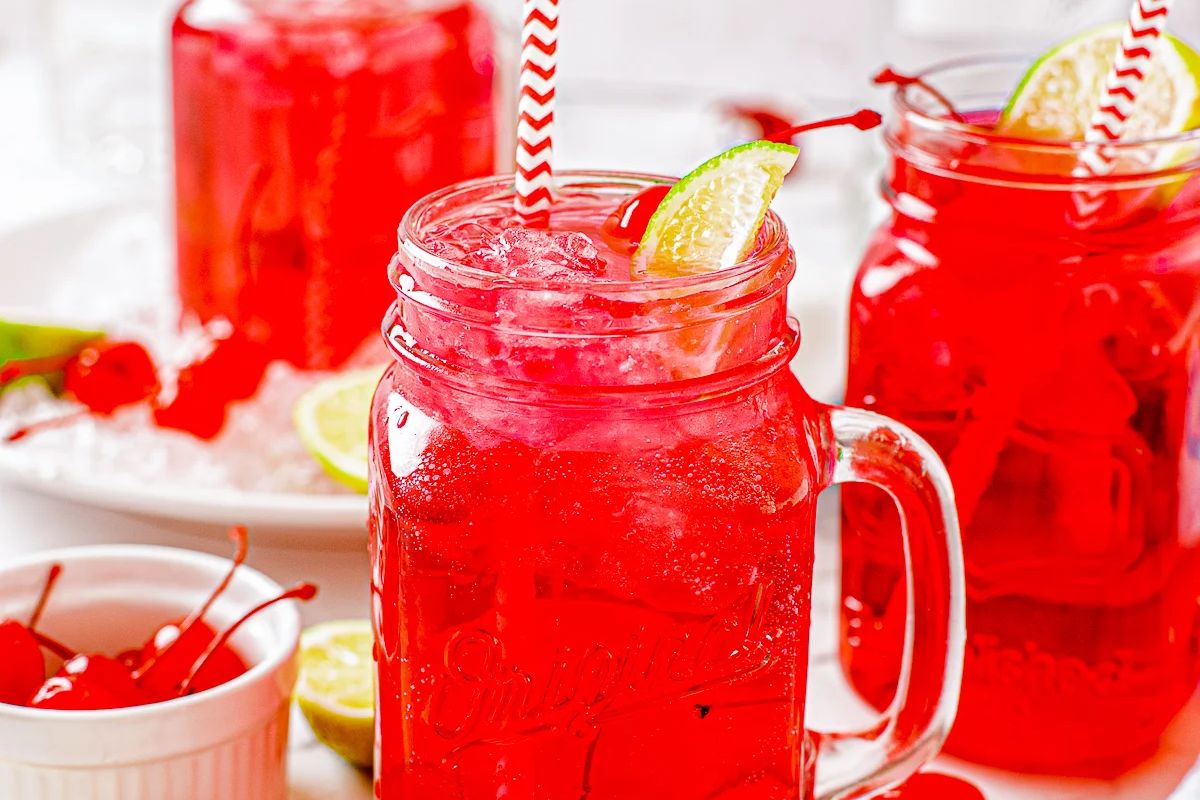 A mason jar filled with red cherry limeade, garnished with a lime wedge and a red and white striped straw, showcases one of the best cherry recipes. Another jar and a bowl of cherries are in the background.