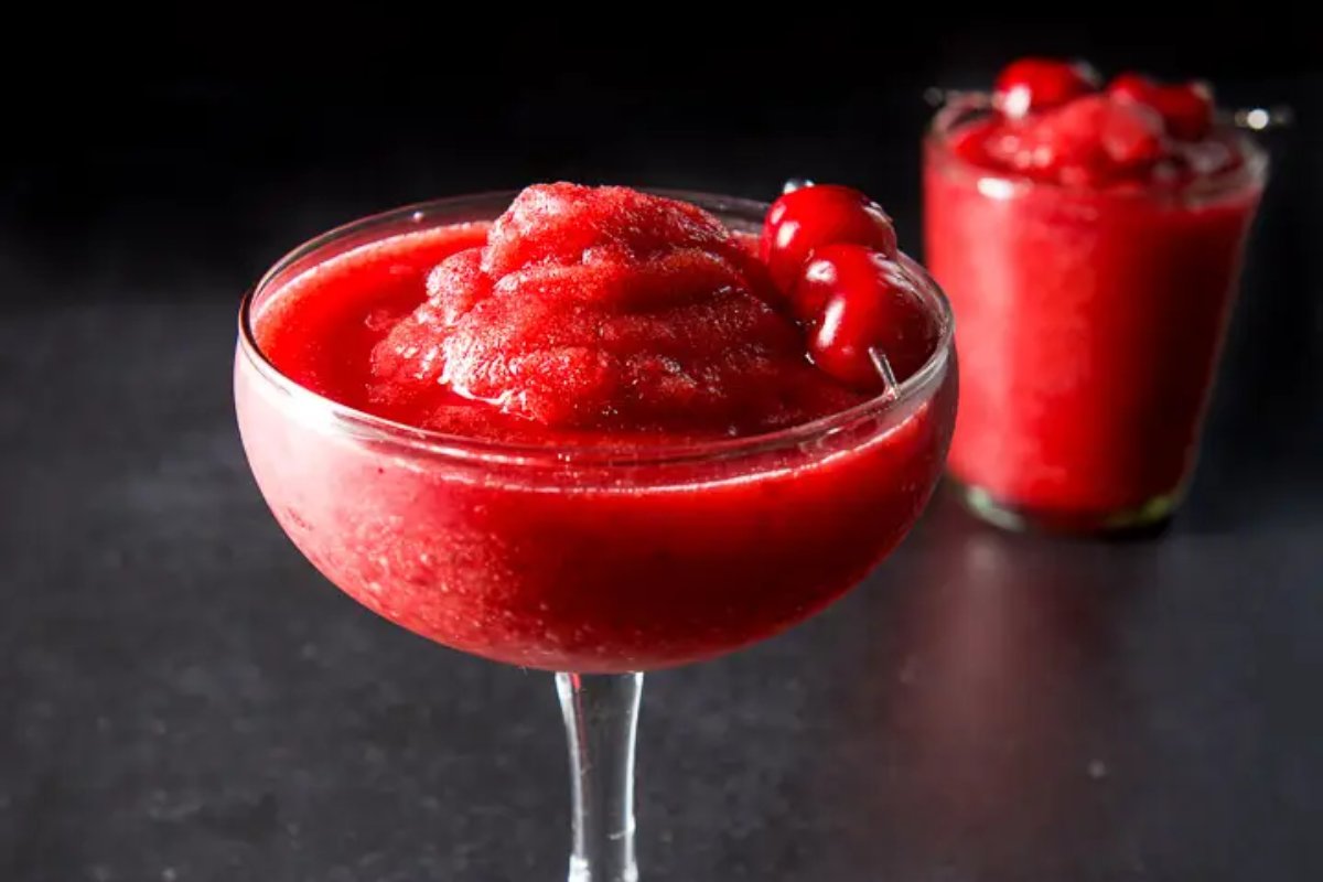 A frosty, cherry-red frozen cocktail in a glass, garnished with two cherries on a skewer. This delightful treat is just one of many tasty cherry recipes you could enjoy. Another similar drink with cherries is blurred in the background.