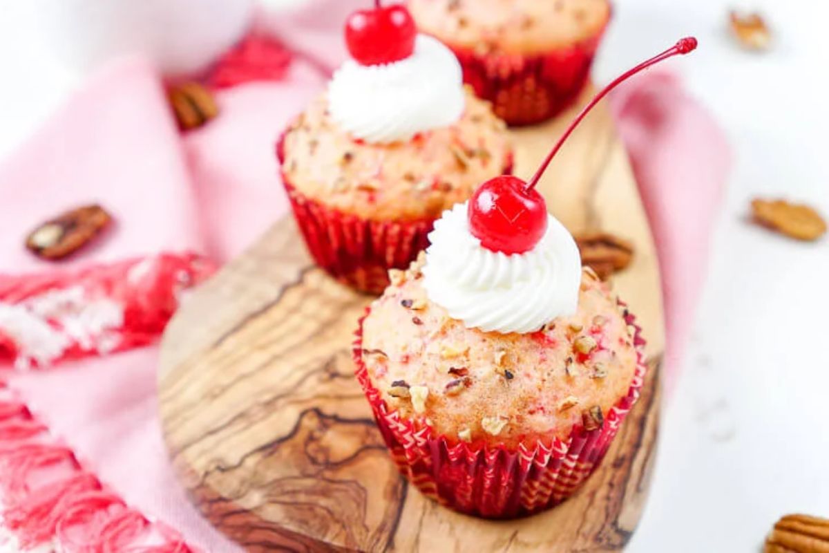 Three cherry-topped cupcakes with white frosting and red sprinkles in red wrappers sit on a wooden board with a pink cloth beside them, perfect for anyone exploring delightful cherry recipes.