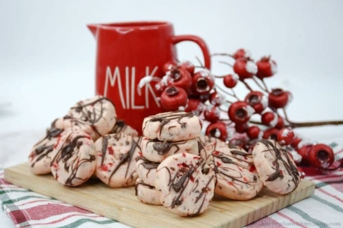 A pile of chocolate-drizzled cookies on a wooden board, placed next to a red pitcher labeled "Milk" and decorative red berries, showcasing our favorite inspiration from cherry recipes.