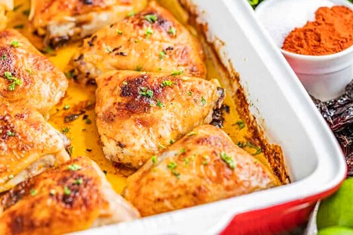 Baked chicken thighs, garnished with parsley, in a rectangular dish. A small bowl of paprika sits nearby, perfect for those exploring new chicken thigh recipes.