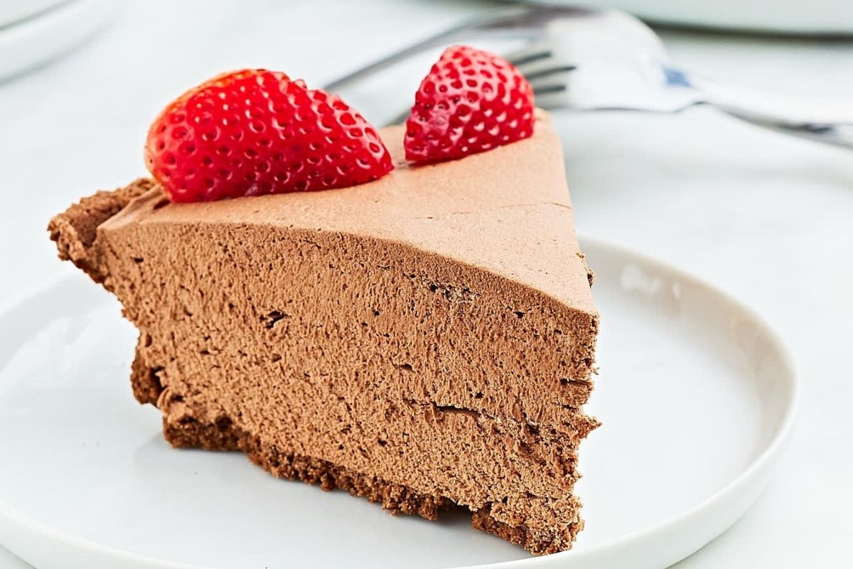 A slice of chocolate mousse pie topped with two strawberry halves on a white plate, with a fork in the background. This is one of those easy desserts that looks as good as it tastes.