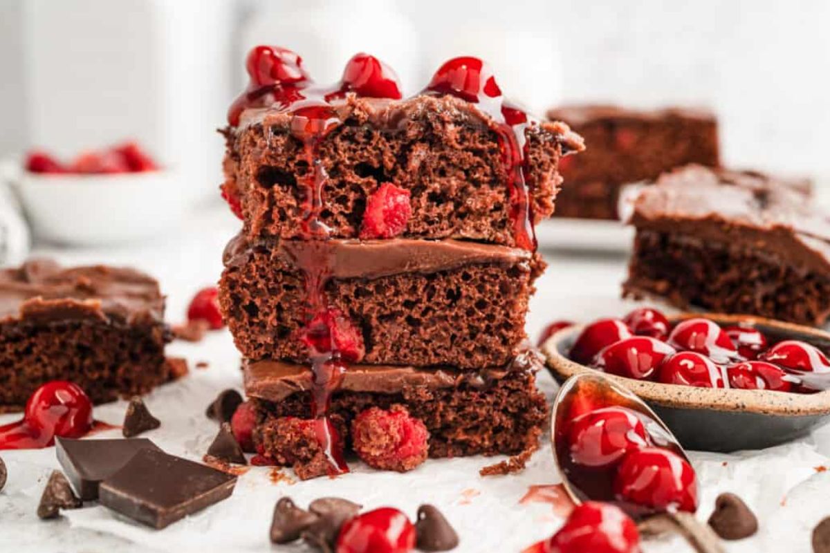 A stack of chocolate cherry cake slices with cherry sauce on top, surrounded by chocolate chunks, a spoon with cherries, and other cake slices on a white surface showcases one of the most delectable cherry recipes you can try.