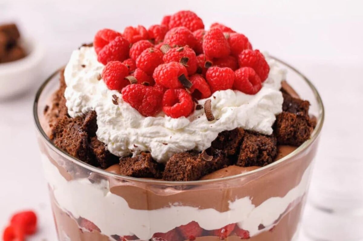 A layered dessert in a glass trifle bowl with chocolate pudding, whipped cream, brownie chunks, and topped with fresh raspberries—a true treat for those who love shortcut desserts.