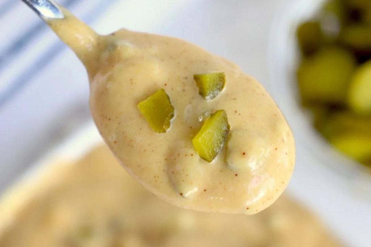 A spoonful of creamy sauce with chopped pickles, inspired by big mac recipes, with a bowl of more pickles blurred in the background.