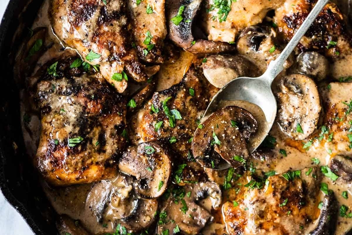 A skillet with chicken thighs and mushrooms in a creamy sauce, garnished with chopped parsley. Perfect for fans of chicken thigh recipes. A spoon rests on top, ready to serve.
