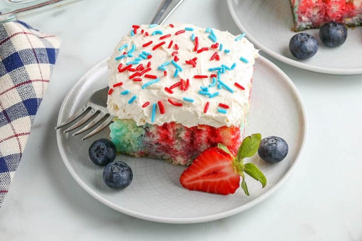 A slice of red, white, and blue poke cake topped with whipped cream and sprinkles sits on a white plate, accompanied by a strawberry and blueberries. This patriotic dessert is complete with a fork perched on the plate's edge.